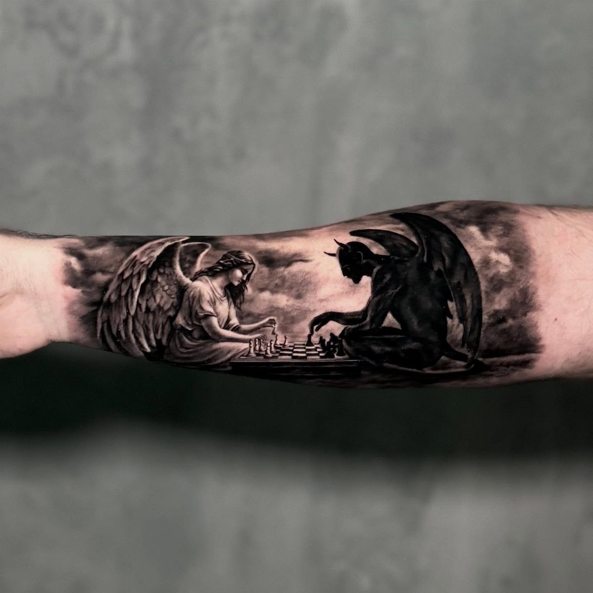 A detailed black and grey tattoo of an angel and a demon playing chess on a forearm.