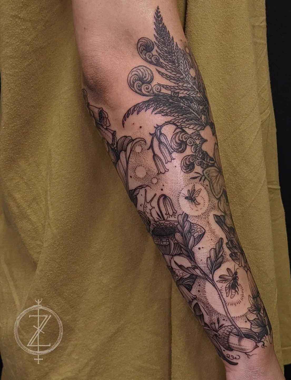 Detailed black and grey botanical tattoo featuring ferns, flowers, and bees on a person's forearm.