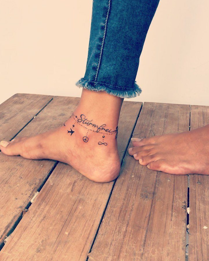 A person's ankle featuring a delicate black ink wrap-around tattoo with symbols and text.