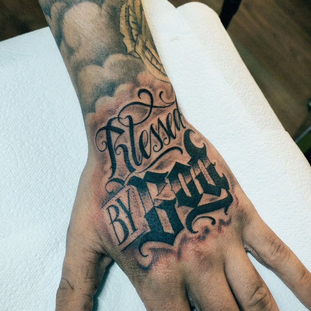 A hand and forearm featuring a large black and grey tattoo reading Blessed BY GOD.