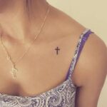 Small black cross tattoo on a person's collarbone with a gold cross necklace.