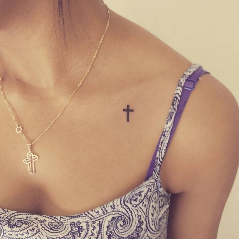 Small black cross tattoo on a person's collarbone with a gold cross necklace.