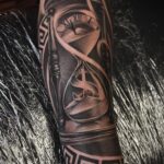 A black and grey realism tattoo of an hourglass with a dollar sign on an arm.