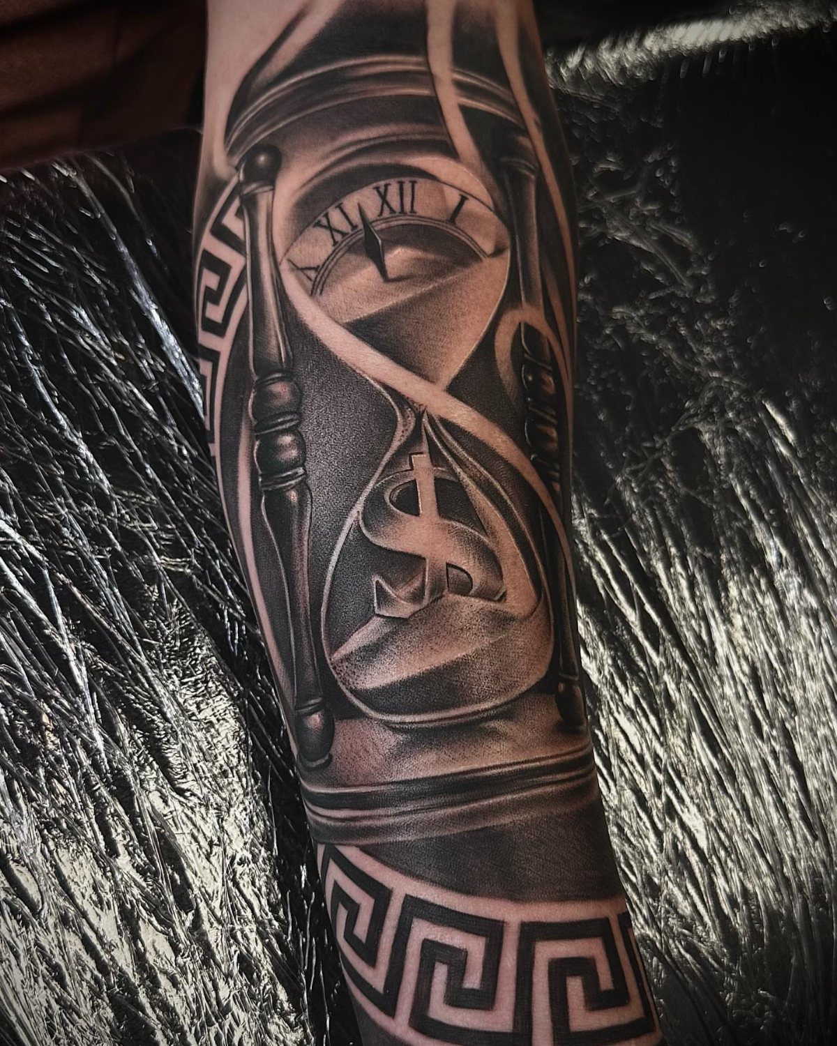 A black and grey realism tattoo of an hourglass with a dollar sign on an arm.