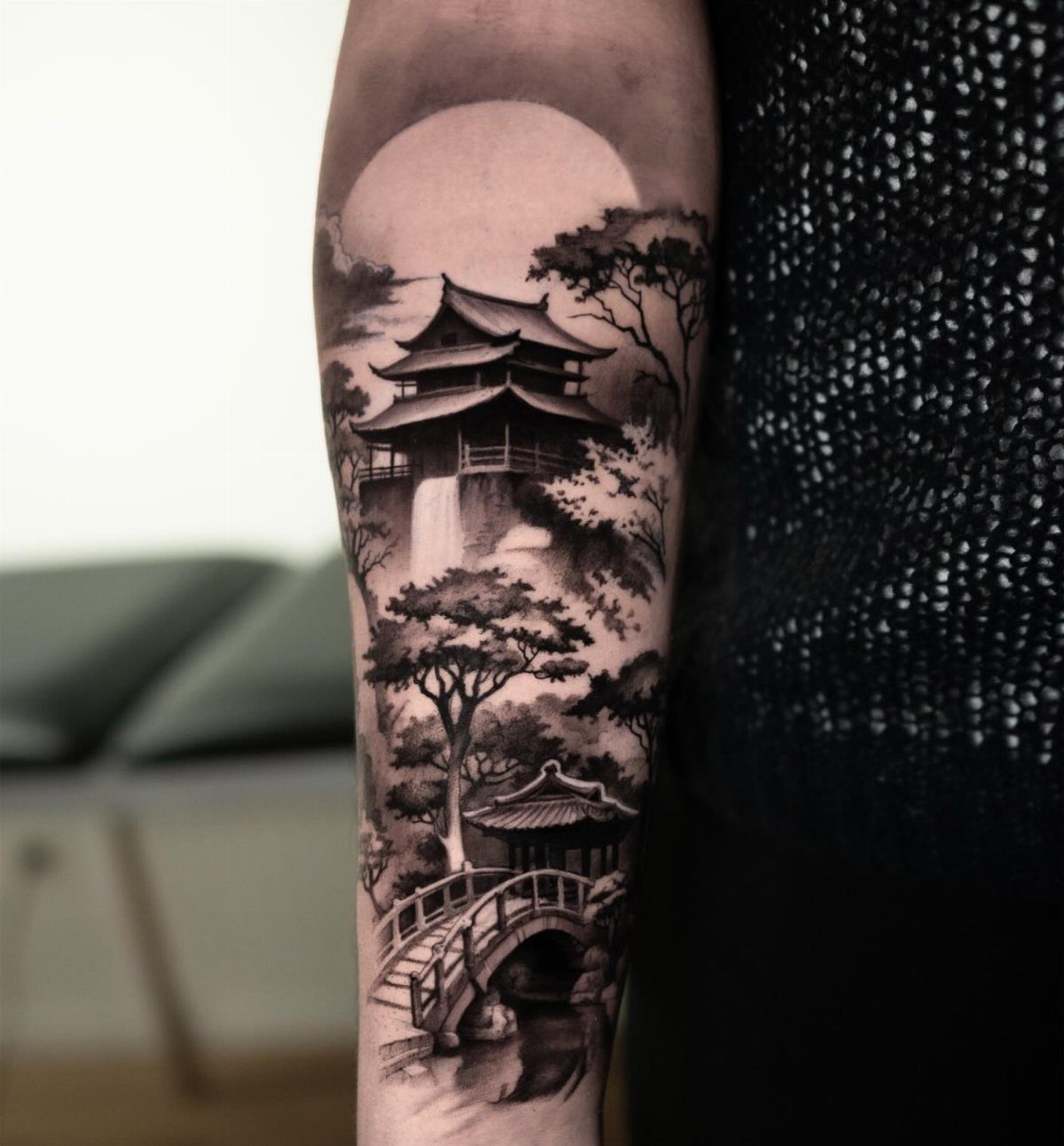 A black and grey tattoo on an arm showing a Japanese landscape with a pagoda, bridge, and sun.