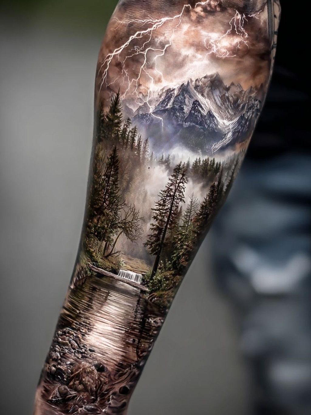 A detailed landscape tattoo on an arm showing mountains, forests, a waterfall, and lightning.