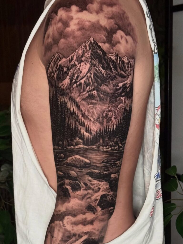 A detailed black and grey landscape tattoo sleeve covering an arm with mountains and forests.