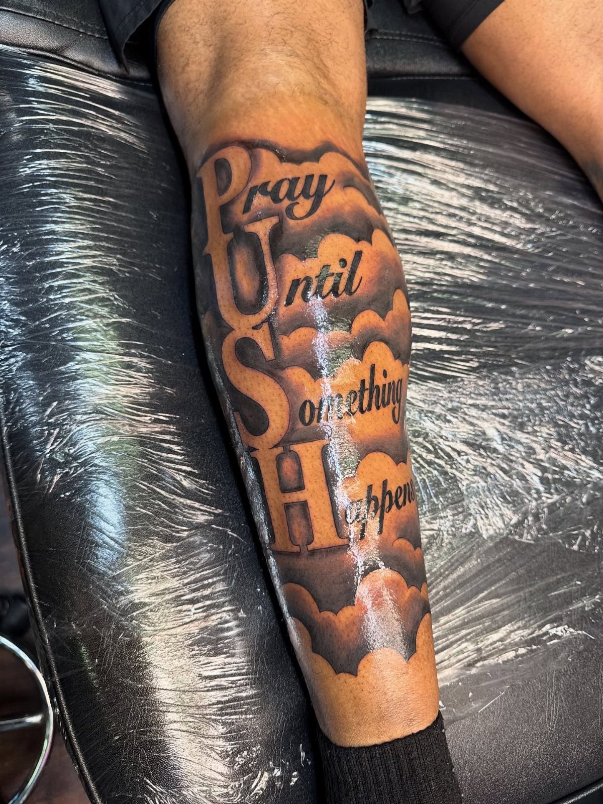 A large black and grey leg tattoo featuring clouds and inspirational text on a person's lower leg.