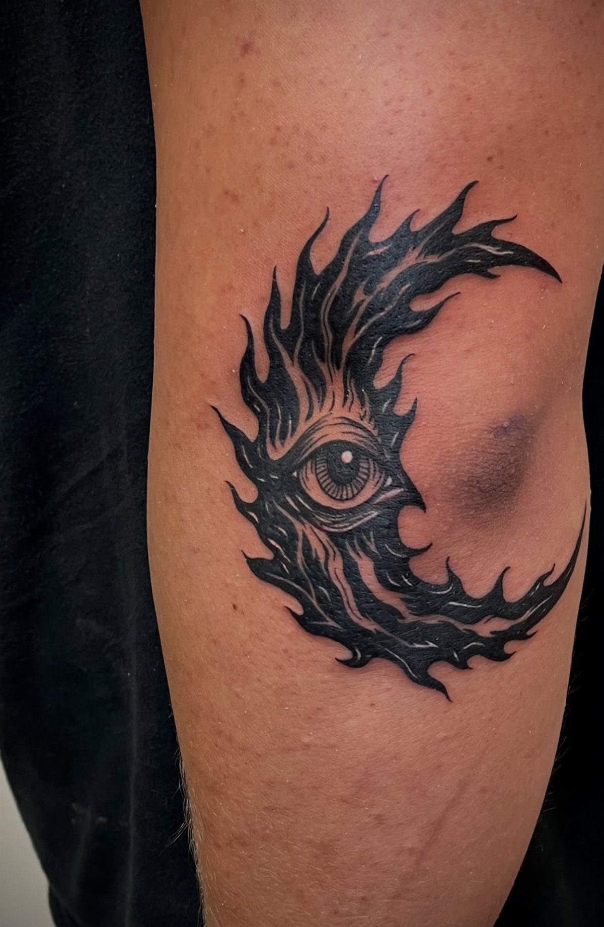 A black ink tattoo of a crescent moon with flames and a central eye on skin.
