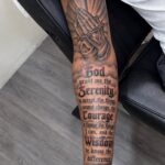 A forearm tattoo depicting praying hands with a rosary and the Serenity Prayer text.