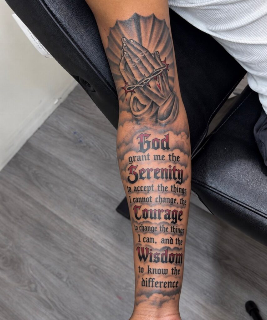 A forearm tattoo depicting praying hands with a rosary and the Serenity Prayer text.