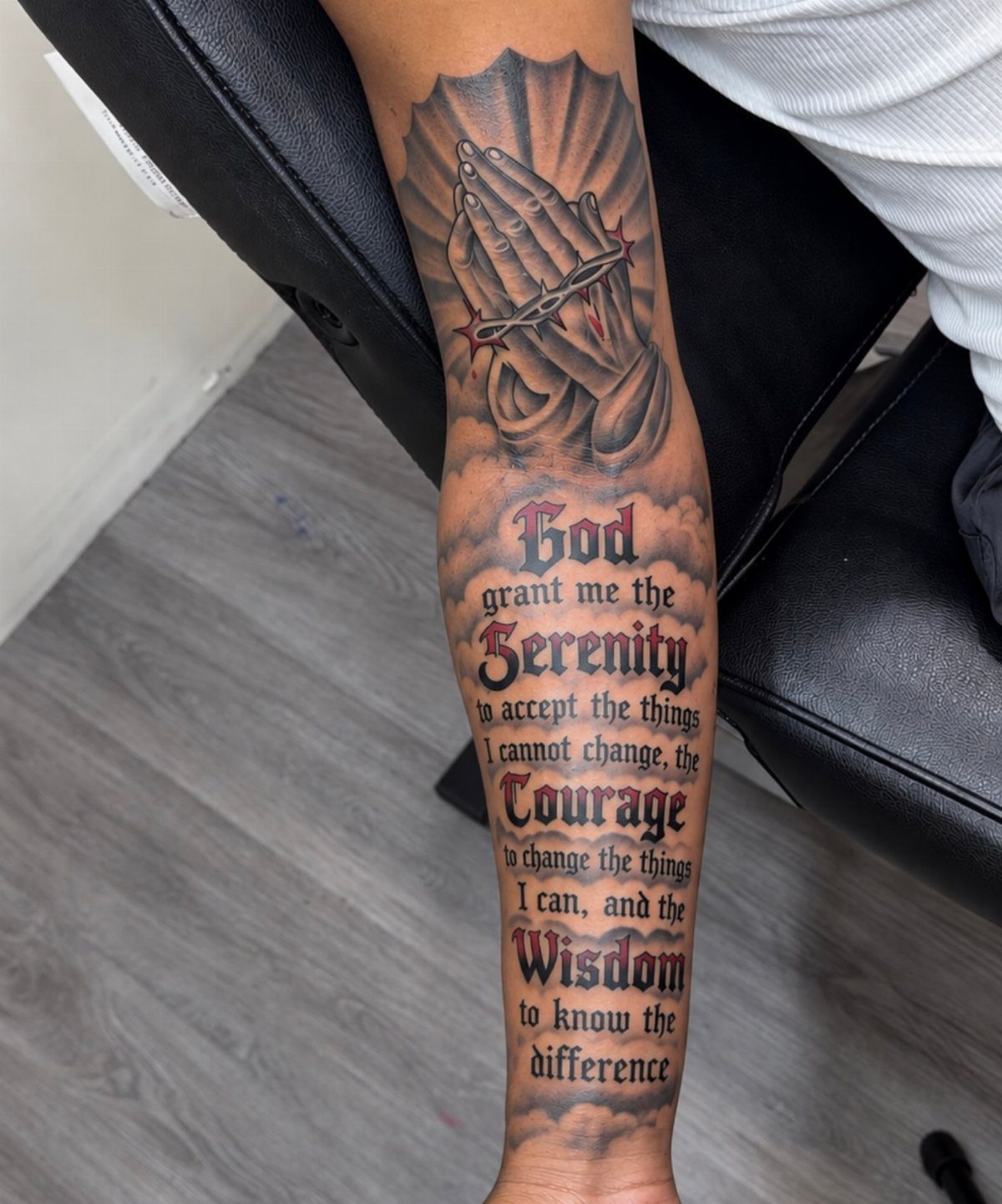 A forearm tattoo depicting praying hands with a rosary and the Serenity Prayer text.