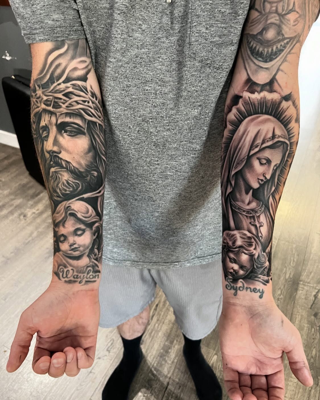 Two tattooed forearms displaying large black and grey religious portraits and names.