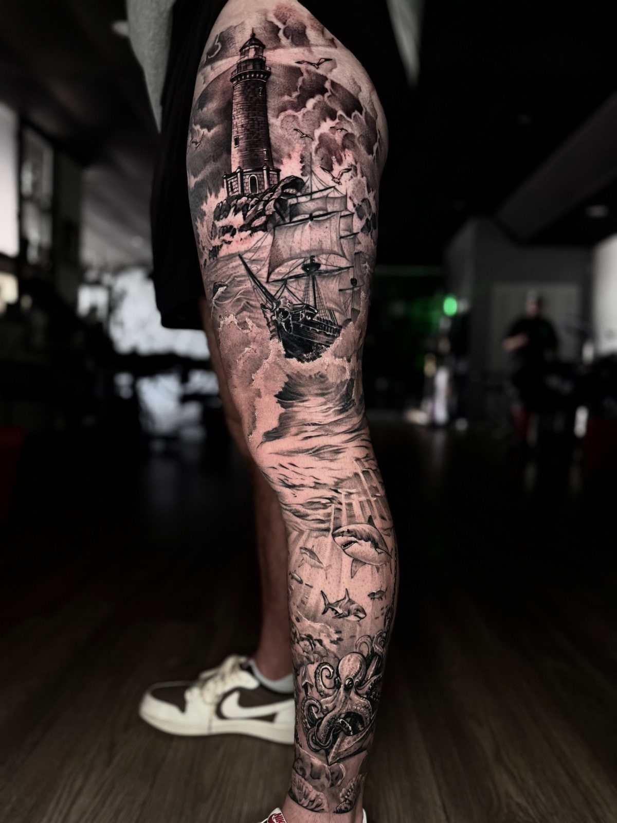 A detailed black and grey leg tattoo depicting a lighthouse, sailing ship, and octopus.