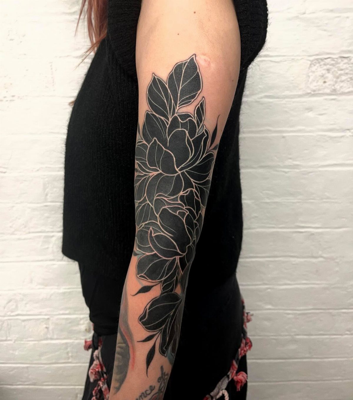 Large black and grey floral tattoo covering a person's arm against a white brick wall.