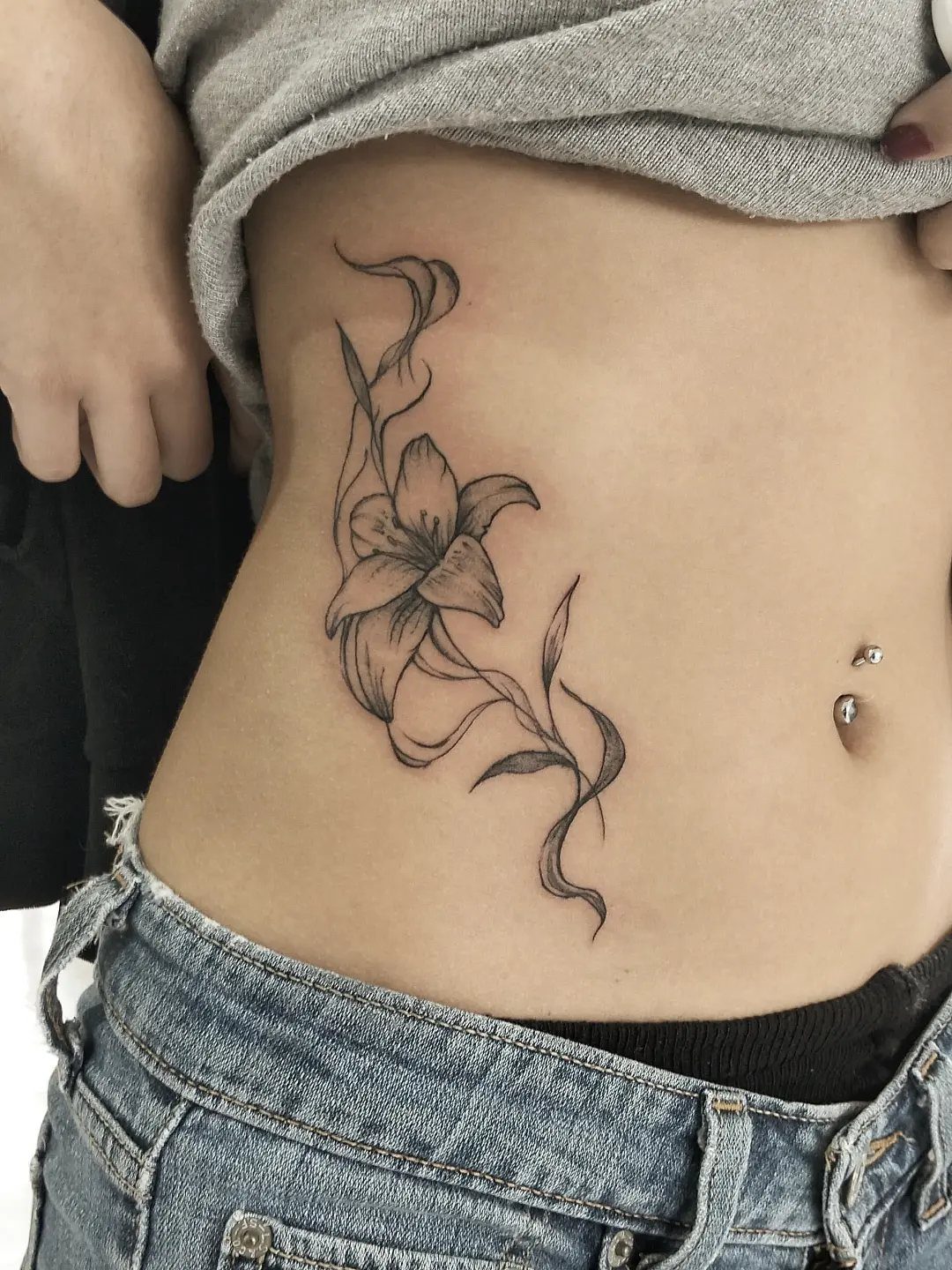 A fine line black ink lily tattoo with flowing stems on a person's abdomen.
